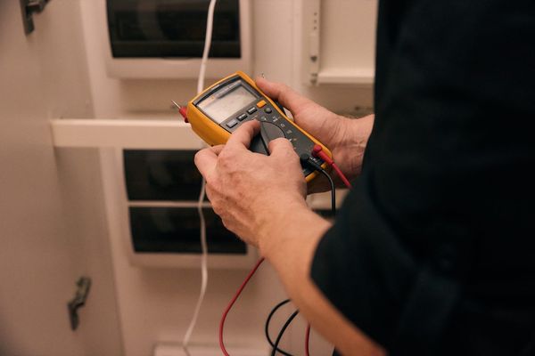 Hands holding a Fluke digital multimeter during electrical fault testing at a residential switchboard