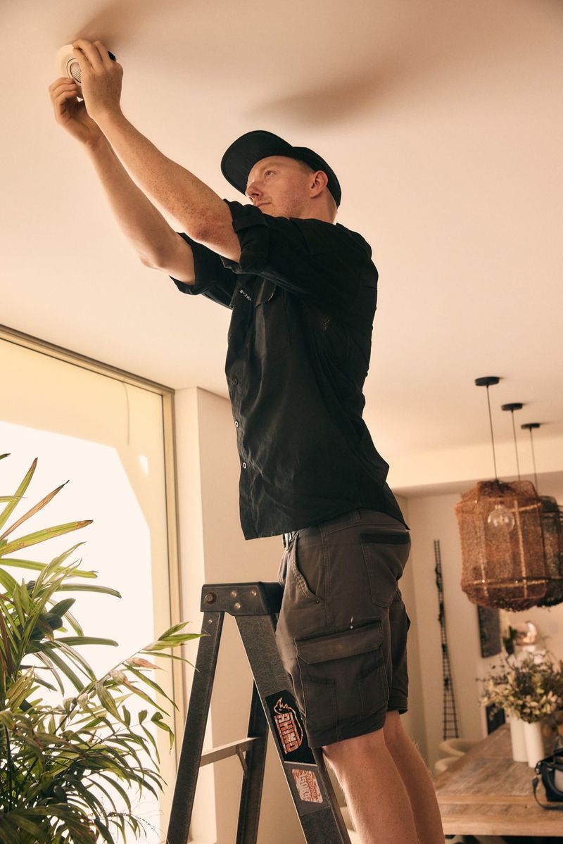 One Out Electrical electrician on a ladder installing a smoke alarm on a ceiling in a Hobart living room